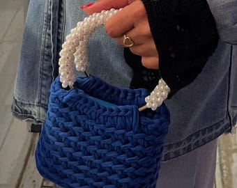 May include: A small, square, royal blue handbag with a woven texture. The bag has a handle made of white pearls. The bag is held by a person wearing a black lace sleeve and a denim jacket.