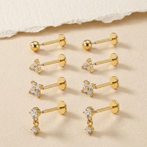 May include: A set of eight gold-tone stud earrings arranged in four pairs. The designs include plain spheres, three-stone clusters, four-stone flower shapes, and dangling two-stone drops, all featuring clear, sparkling stones on flat-back posts.