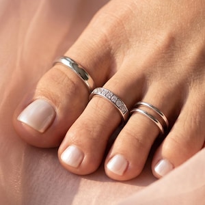 May include: A set of three silver-toned toe rings worn on the toes. One ring is a plain polished band, the second is a channel-set band featuring small clear stones, and the third is a double-banded ring with a smooth, minimalist finish.