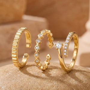 May include: Three gold-toned rings with open designs. The first ring features a ribbed texture, the second has a geometric pattern with small clear stones, and the third is a bypass ring with a single larger stone and a row of smaller clear stones.