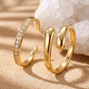 May include: Two gold-toned open rings. One ring features a thin band encrusted with a row of small, clear, sparkling stones. The second ring has a smooth, polished, solid gold-toned surface with a rounded, teardrop-shaped end design.