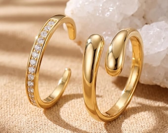 Könnte beinhalten: Zwei goldfarbene offene Ringe. Ein Ring hat ein schmales Band, das mit einer Reihe kleiner, klarer, funkelnder Steine besetzt ist. Der zweite Ring hat eine glatte, polierte, goldfarbene Oberfläche mit einem abgerundeten, tropfenförmigen Design.