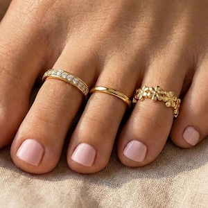 May include: Close-up of a foot with three gold toe rings. One ring is a band with small clear stones, another is a simple band, and the third features a floral design. The toenails are painted a light pink color.