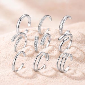 Könnte beinhalten: Eine Sammlung von neun silberfarbenen offenen Ringen mit verschiedenen Designs, darunter glatte Bänder, Bänder mit eingelassenen klaren Steinen, strukturierte Muster und mehrlagige Stile aus poliertem Metall.