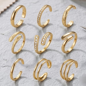 Könnte beinhalten: Neun goldfarbene Zehenringe in verschiedenen Designs werden auf einer hellgrauen Oberfläche präsentiert. Die Ringe umfassen schlichte Bänder, Bänder mit kleinen, klaren Steinen und strukturierte Muster. Einige Ringe haben ein offenes Design.