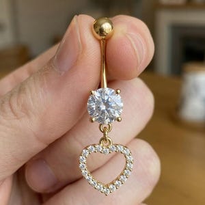 May include: A gold-colored belly button ring featuring a large, clear, round gemstone and a dangling heart-shaped charm. The heart is outlined with small, sparkling stones. The top of the ring has a small, round ball.