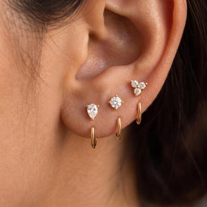 May include: Three gold earrings worn in a row on an earlobe. The first is a pear-shaped stud, the second is a round solitaire stud, and the third is a three-stone cluster stud. Each stud is paired with a small, plain gold hoop earring underneath.