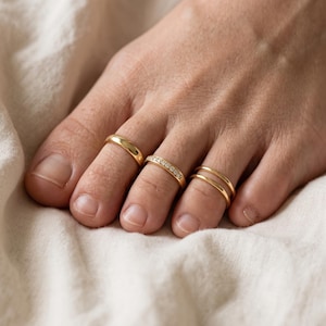May include: Three gold-toned toe rings worn on the toes. The first is a plain polished band, the second features a row of small clear stones, and the third is a double-banded design. Each ring is crafted from a smooth, reflective metal.