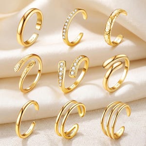 May include: Nine gold-colored rings in various designs are displayed on a white surface. The rings include open-ended bands, some with clear gemstones, and others with textured or double-banded designs. Each ring is marked with "14K GF".