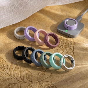 May include: A collection of silicone rings in various colors, including gray, purple, pink, and burgundy, alongside black, blue, and green. A silver ring and a charging base are also present. The rings are arranged on a textured surface.