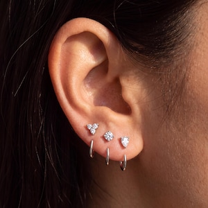 May include: Three silver-toned earrings worn in a row on an earlobe. Each earring features a small silver hoop base with a distinct decorative stud attachment: a three-stone cluster, a single round brilliant-cut stone, and a pear-shaped stone.