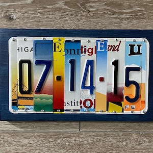 May include: A blue wooden frame with a date made from license plates. The date is 07.14.15. The license plates are from different states and have various colors and designs.