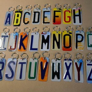 May include: A set of 26 alphabet letter keychains made from recycled license plates. Each letter is a different color and has a unique design. The keychains are attached to silver key rings.