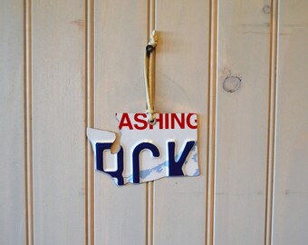 May include: A blue and white metal ornament shaped like the state of Washington. The ornament has the word "WASHING" printed in red letters and the letters "OCK" printed in blue letters. The ornament is hanging from a string.