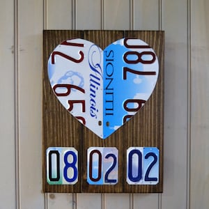 May include: A wooden heart-shaped sign with a blue and white Illinois license plate on the left side and a red and white license plate on the right side. The date 08 02 02 is displayed below the heart.