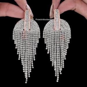 May include: A pair of dazzling, silver-colored rhinestone earrings. Each earring features a half-circle top with a vertical bar and a cascade of dangling rhinestones. The earrings are set against a black background.