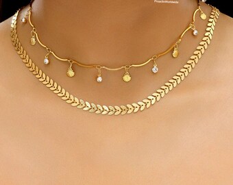 May include: Two gold necklaces layered on a person. The top necklace has a wavy design with small seashell charms and clear crystals. The bottom necklace has a leaf-like design. The necklaces are on a person's neck.
