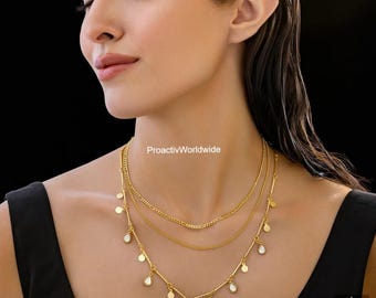 May include: A layered gold necklace set. The top chain is a simple curb chain, the middle chain is a delicate cable chain, and the bottom chain features small, pear-shaped pendants. The necklace is worn against a black background.
