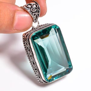 Pode incluir: Um pingente retangular com uma grande pedra preciosa facetada de cor verde-azulado. A pedra é engastada em uma moldura de metal prateado com detalhes ornamentados e uma argola decorativa.