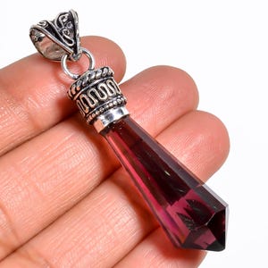 May include: A faceted, elongated pendant with a deep red gemstone. The pendant has a silver-colored metal cap with ornate detailing and a decorative bail. The gemstone is a rich, transparent color, and the pendant is held in a hand.
