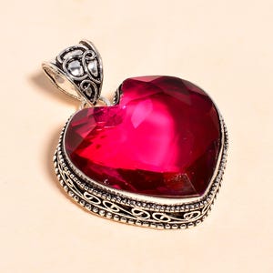 Op de afbeelding: Een hartvormige hanger met een grote, gefacetteerde, dieprode edelsteen. De edelsteen is gezet in een zilverkleurig metalen frame met een sierlijk ontwerp. De hanger heeft een decoratieve beugel.