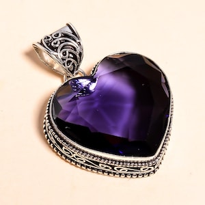 Op de afbeelding: Een hartvormige hanger met een grote, facetgeslepen, dieppaarse edelsteen. De edelsteen is gezet in een zilverkleurig metalen frame met sierlijke details. De balein, waar een ketting aan zou worden bevestigd, heeft ook decoratieve krullen.