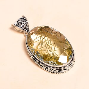 May include: Oval-shaped pendant featuring a large, translucent yellow gemstone with golden, needle-like inclusions. The gemstone is set in a decorative silver-colored metal frame with an ornate bail.