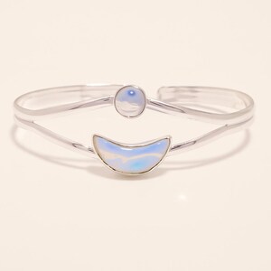 Peut inclure: Un bracelet manchette argenté orné d'un croissant de lune et d'une pierre circulaire. La lune et le cercle sont bleu clair et blancs. Le bracelet a un design minimaliste, probablement en argent sterling.