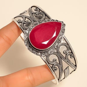 Puede incluir: Una pulsera de plata con un diseño ornamental y ondulado. La pulsera presenta una gran gema roja en forma de lágrima en el centro, rodeada por un borde detallado con cuentas. La pulsera está sujeta por una mano.
