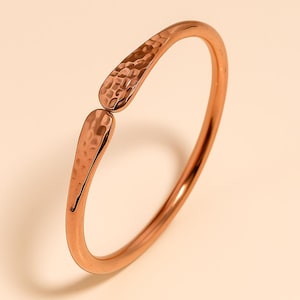 Puede incluir: Un brazalete de puño abierto de color cobre. El brazalete presenta una textura martillada en los dos extremos decorativos. El brazalete es de diseño sencillo y elegante.