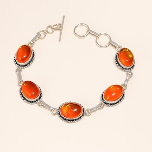 Puede incluir: Un brazalete de plata con piedras preciosas ovales de color naranja. Cada piedra está engastada en un bisel de plata con un diseño de cuerda trenzada. El brazalete tiene un cierre de palanca. Las piedras tienen un aspecto cálido, similar al ámbar. Mide unos 19 cm.