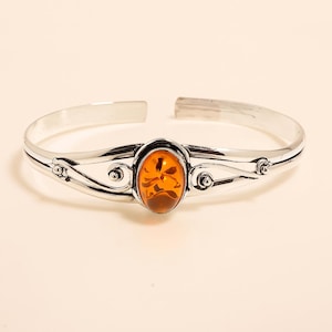 May include: A silver cuff bracelet featuring an oval amber stone at the center. The bracelet has decorative swirls and small circular accents on either side of the amber. The amber stone is a warm orange color.