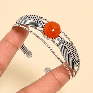 Pode incluir: Uma pulseira de punho em tom prateado com uma pedra preciosa laranja, redonda e lisa, engastada num bisel recortado. A banda é composta por vários fios finos texturizados e é acentuada com dois motivos de folhas prateadas com nervuras gravadas.