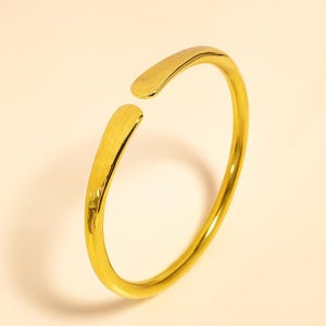 Puede incluir: Un brazalete de puño abierto de color dorado. El brazalete tiene una textura martillada en los extremos y un acabado liso y pulido en la banda curva. El brazalete es de diseño simple y elegante.