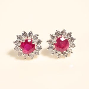 Peut inclure: Deux boucles d'oreilles à clous avec une pierre précieuse rouge foncé ronde au centre, entourée d'un halo de petites pierres précieuses claires. Les boucles d'oreilles ont un design floral, les pierres claires étant disposées comme des pétales.