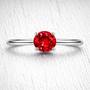 May include: A silver ring featuring a large, round, vibrant red gemstone. The gemstone is held in place by four prongs, and the band is smooth and polished. The ring is set against a white background.