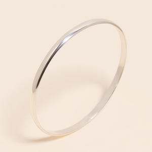 Peut inclure: Un bracelet jonc argenté avec une finition lisse et polie. Le bracelet a un design simple et minimaliste et est de forme circulaire. Le métal reflète la lumière, créant des reflets et des ombres.