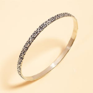 Pode incluir: Uma pulseira de bracelete prateada com um padrão floral. A pulseira tem um interior liso e polido e um exterior texturizado com desenhos repetidos de flores e folhas. A pulseira é circular e parece ser feita de metal.