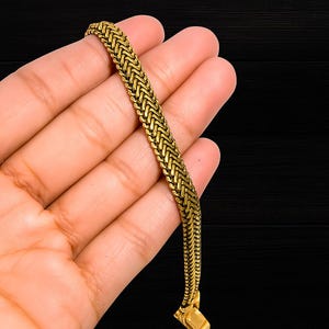 Puede incluir: Una pulsera dorada con un diseño tejido. La pulsera se sostiene en una mano sobre un fondo negro. La pulsera tiene un cierre rectangular.