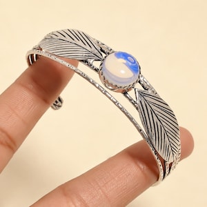Puede incluir: Una pulsera tipo brazalete de tono plateado con dos detalles metálicos en forma de hoja con grabados. Una piedra cabujón redonda, lisa, de color blanco lechoso y azul, está engastada en el centro entre las hojas con un borde festoneado.