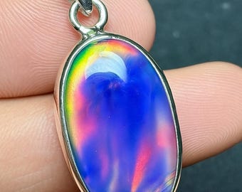 May include: Oval-shaped pendant with a vibrant, iridescent gemstone. The stone displays a spectrum of colors, including blues, purples, and hints of red and yellow. The pendant is set in a silver-colored metal with a loop for a chain.
