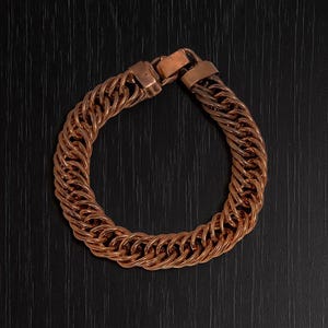 Op de afbeelding: Een koperkleurige armband met een schakelkettingontwerp. De armband is gemaakt van meerdere rijen in elkaar grijpende schakels, waardoor een dikke, getextureerde uitstraling ontstaat. De sluiting heeft een eenvoudig, rechthoekig ontwerp. De armband wordt op een donkere ondergrond getoond.