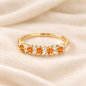 Op de afbeelding: Een gouden ring met een rij afwisselende ronde oranje edelstenen en kleinere heldere witte stenen. De band is van gepolijst goud en de stenen zijn in een doorlopende lijn op de bovenste helft van de ring gezet voor een sprankelend effect.