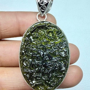 May include: Oval pendant featuring a textured, olive-green stone set in a silver-coloured metal frame. The pendant has a decorative bail with a swirling design. The stone's surface has a unique, organic appearance.