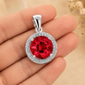 May include: A circular pendant featuring a large, round, vibrant red gemstone at the centre, surrounded by a halo of small, clear, sparkling white stones set in a polished silver-toned metal frame with a matching silver-toned bail at the top.