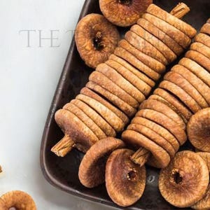 Puede incluir: Una bandeja rectangular marrón oscuro llena de higos secos. Los higos están dispuestos en pilas e individualmente, mostrando su color marrón natural y superficie texturizada. La imagen está tomada desde arriba sobre un fondo blanco.