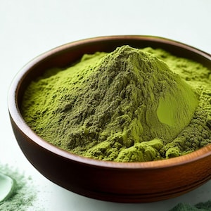 May include: A wooden bowl brimming with bright green powder, likely a spice or herb. The powder is finely ground and has a slightly textured appearance. The bowl is set against a light background, with a few scattered granules of the green powder nearby. The text "THE DESI SPICES" is at the bottom.