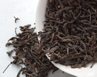 May include: A white, slightly tilted bowl overflowing with dark brown loose leaf tea. The tea spills out onto a white surface, creating a textured contrast. The image is well-lit, highlighting the tea's rich color and detail.