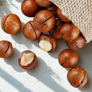 Puede incluir: Primer plano de nueces de macadamia marrones, algunas enteras y otras agrietadas, saliendo de un saco de yute. Las nueces están esparcidas sobre una superficie blanca, con una nuez que revela su interior cremoso.