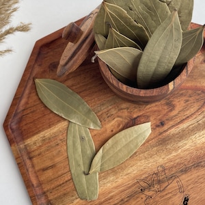 May include: A wooden serving tray with a hexagonal shape holds a small wooden bowl filled with dried bay leaves. Several bay leaves are scattered on the tray. The tray has a logo etched on it. The leaves are a muted green color.